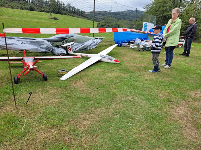Ein weißer Segler und ein rotes Modellflugzeug liegen auf einer Wiese bei einem Flugplatzfest.