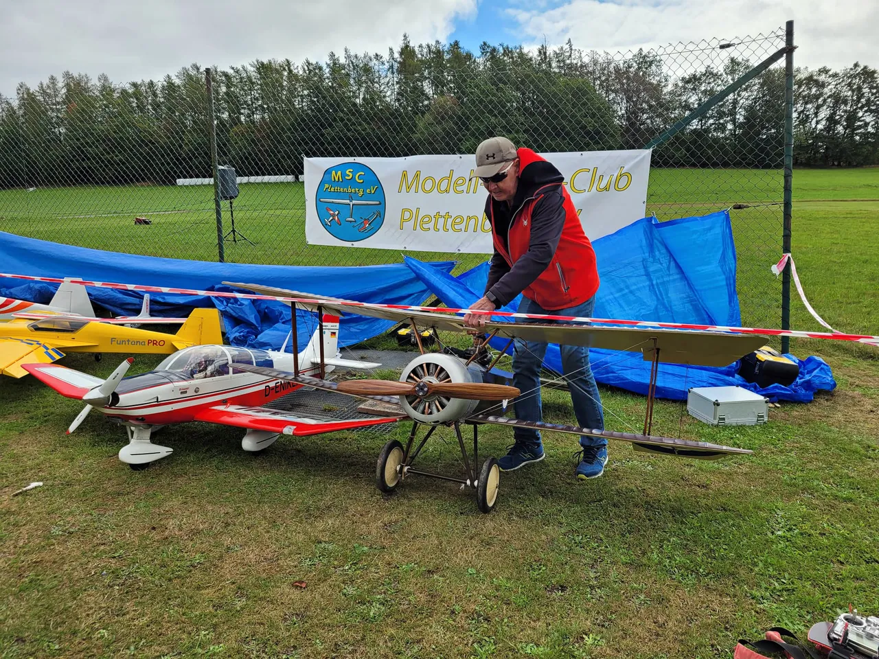 Ein Mann in einer roten Weste arbeitet an einem großen Modellflugzeug auf einer Wiese vor einem Banner des MSC Plettenberg.