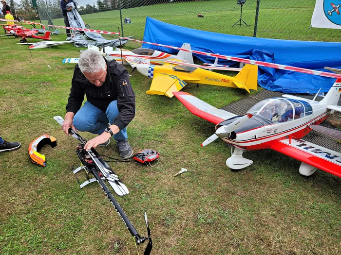 Ein Mann kniet auf einer Wiese und arbeitet an einem großen ferngesteuerten Modellflugzeug, während im Hintergrund weitere Modelle liegen.