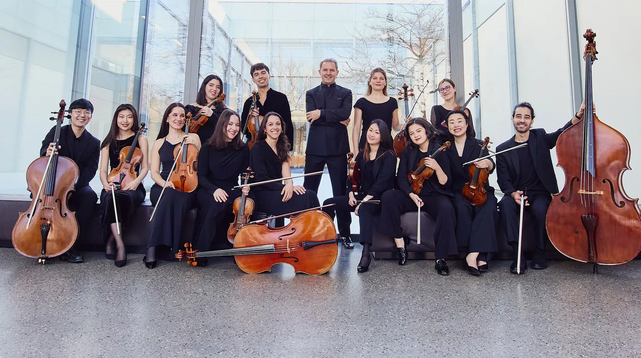 Ein Ensemble von 15 Musikerinnen und Musikern, die Streichinstrumente spielen, posieren für ein Gruppenfoto. Sie tragen größtenteils schwarze Kleidung und befinden sich in einem hellen Raum mit großen Fenstern.