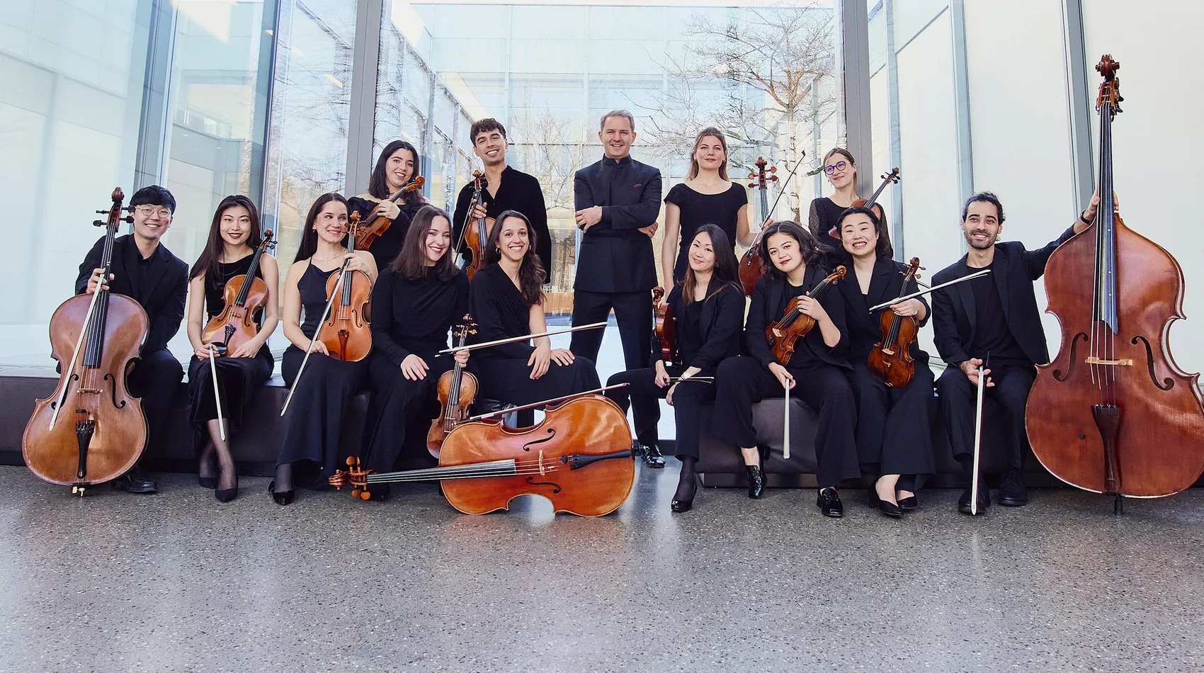 Ein Ensemble von 15 Musikerinnen und Musikern, die Streichinstrumente spielen, posieren für ein Gruppenfoto. Sie tragen größtenteils schwarze Kleidung und befinden sich in einem hellen Raum mit großen Fenstern.