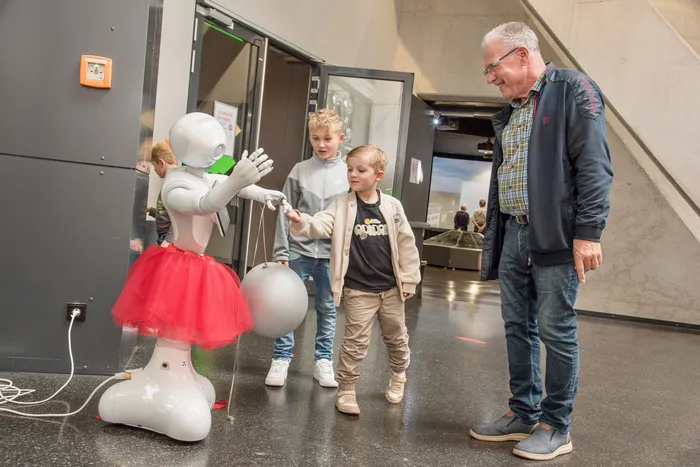 Ein weißer Roboter mit rotem Tutu hält eine graue Kugel und interagiert mit zwei Jungen und einem Mann in einem Museum.