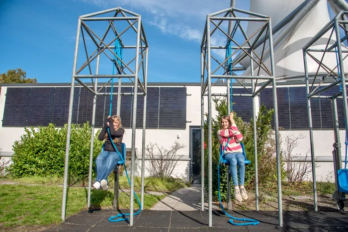 Zwei Mädchen sitzen auf blauen Seilschwingen in hohen Metallrahmen vor einem Gebäude mit Solarmodulen.