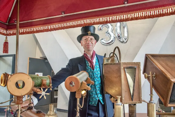 Ein Mann mit Zylinder und gemusterter Weste steht hinter historischen wissenschaftlichen Geräten vor einem roten Baldachin und einem silbernen „30“-Luftballon.