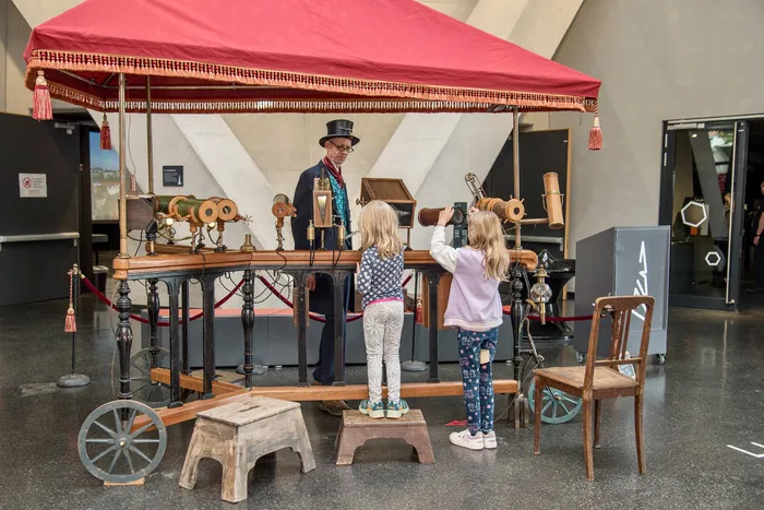 Ein Mann im Zylinder steht an einem hölzernen Stand unter einem roten Baldachin, während zwei Kinder durch darauf montierte Fernrohre blicken.