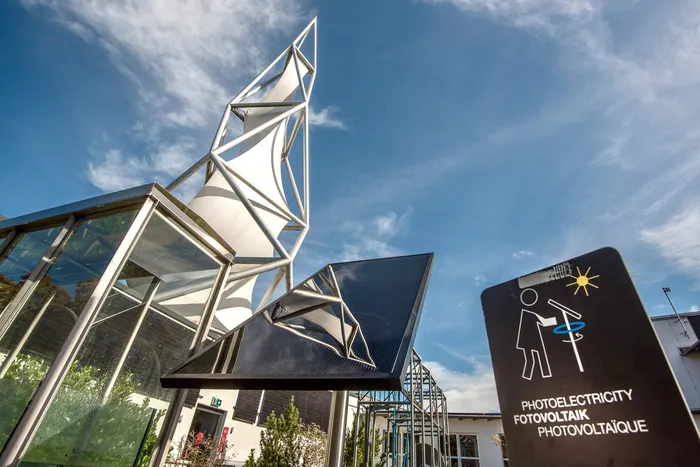 Weiße, segelartige Architektur unter blauem Himmel, ein Solarpanel und ein Hinweisschild mit der Aufschrift „PHOTOELECTRICITY FOTOVOLTAIK PHOTOVOLTAÏQUE“.