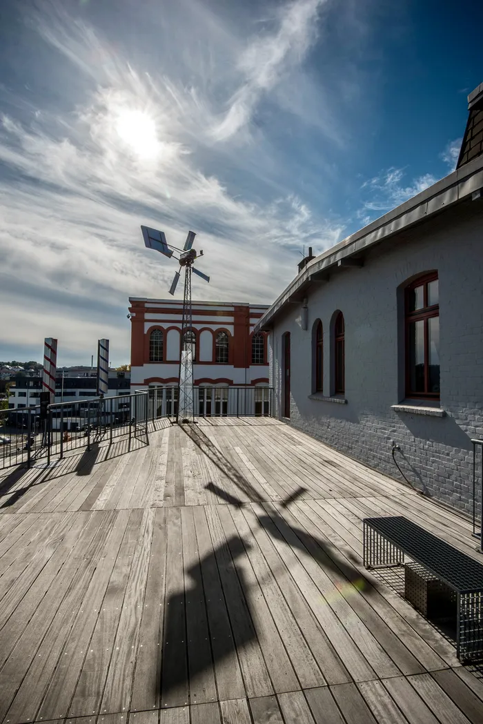 Holzterrasse vor einem hellgrauen Gebäude mit einem hohen Metallmast, der einen langen Schatten wirft, vor einem roten Gebäude unter blauem Himmel.