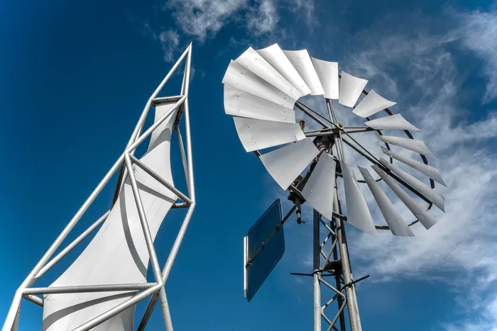 Eine weiße, segelartige Struktur und eine silberne Windturbine vor blauem Himmel mit Wolken.