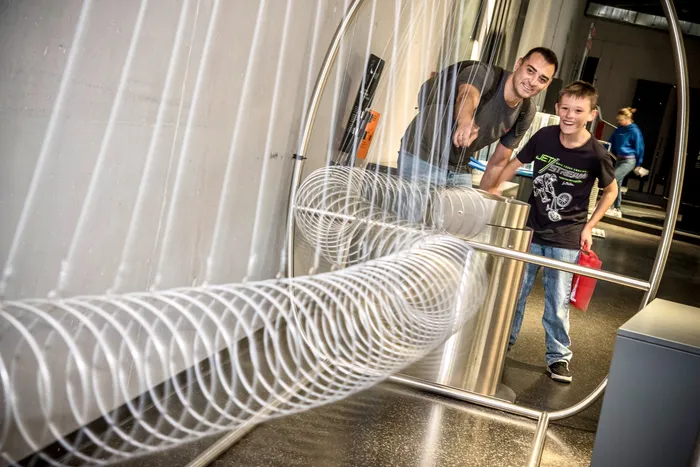 Ein Mann und ein Junge interagieren lachend mit einer optischen Installation aus weißen, spiralförmigen Ringen in einem Museum.