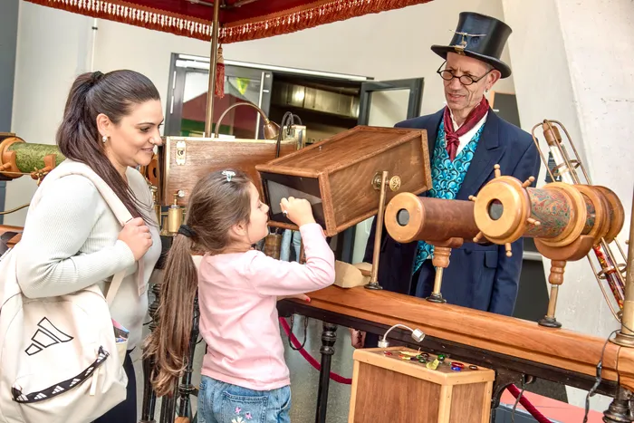 Ein Mädchen blickt durch ein hölzernes optisches Gerät an einem Stand im Museum Phänomenta, begleitet von einer Frau und einem Mann mit Zylinder.