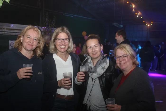 Vier lächelnde Frauen halten Plastikbecher mit Getränken bei einem Scheunenfest.
