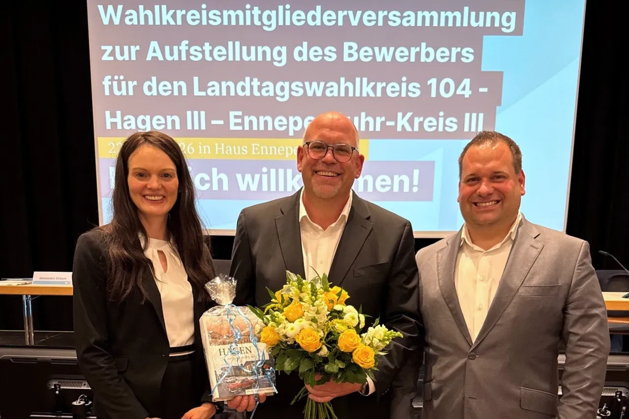 Veit Kuhl hält einen Blumenstrauß und eine Urkunde, flankiert von Anna Dern links und Dennis Rehbein rechts. Im Hintergrund ein Banner mit dem Aufdruck zur Aufstellung des Landtagskandidaten für Wahlkreis 104.