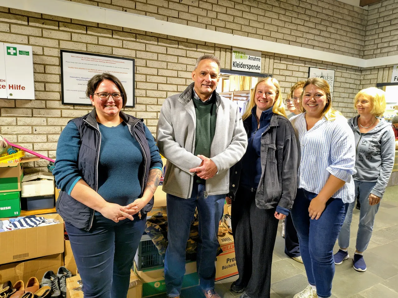 Fünf Personen stehen vor Kisten mit Spenden und geben sich die Hände. Im Hintergrund ein Schild mit der Aufschrift „Kleiderspende“.