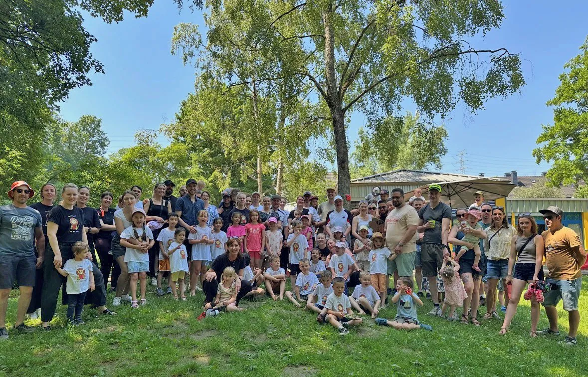 Eine große Gruppe von Erwachsenen und Kindern steht lächelnd für ein Gruppenfoto zusammen. Im Hintergrund sind Bäume und ein Gebäude zu sehen.