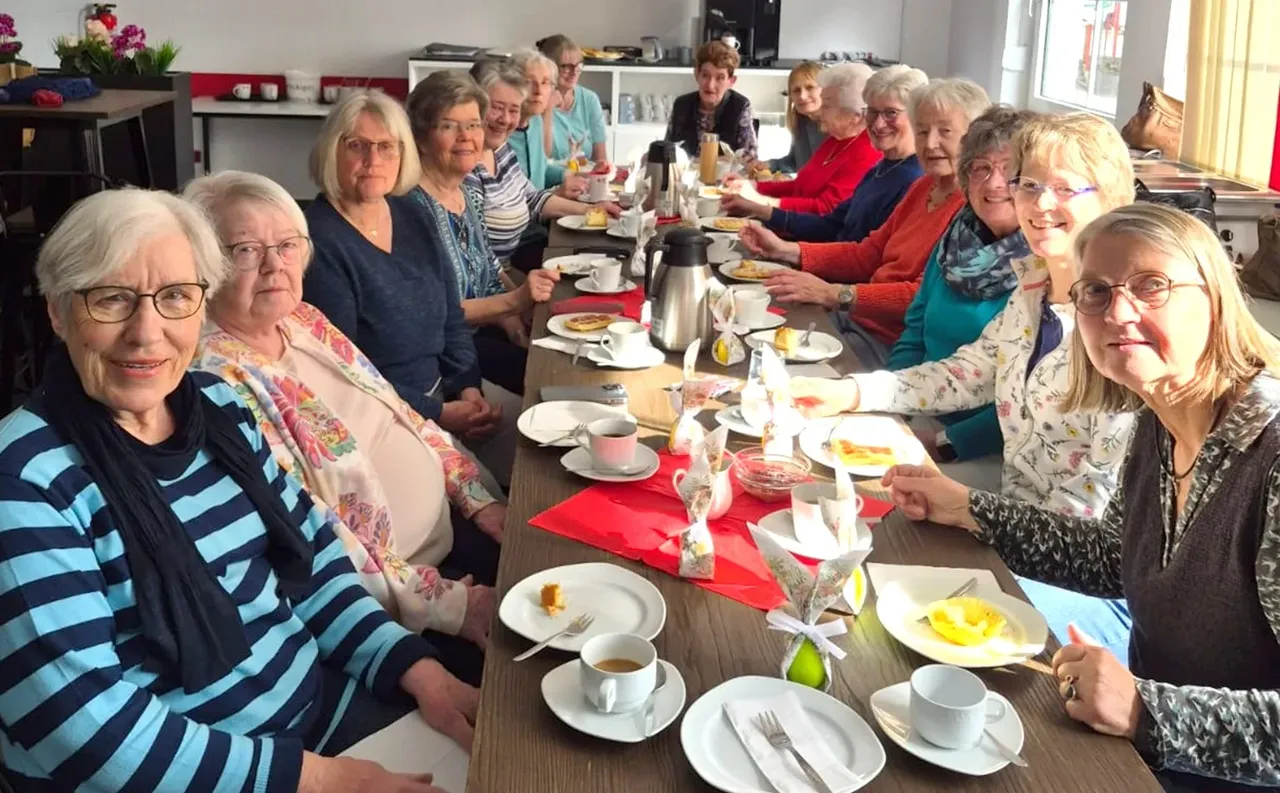 Eine Gruppe älterer Frauen sitzt an einem langen Tisch und isst Kuchen, während sie sich unterhalten. Auf dem Tisch stehen Tassen, Teller mit Kuchenstücken und Besteck.