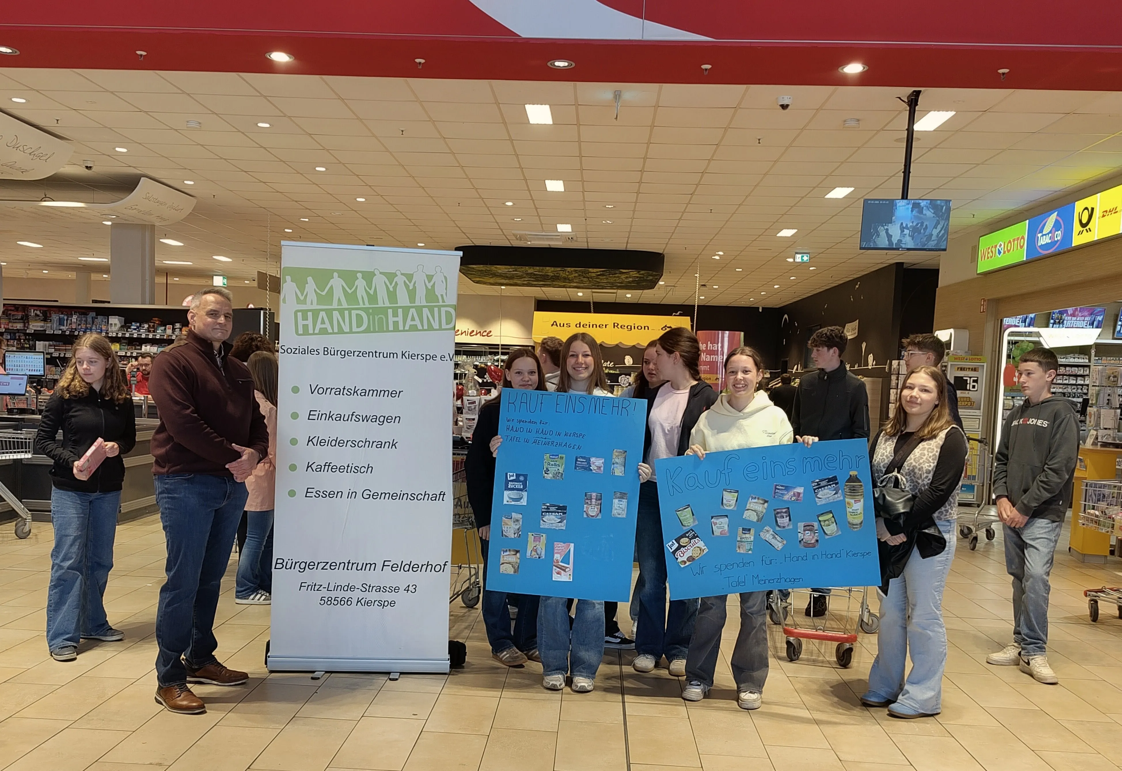 Eine Gruppe von Menschen steht in einem Supermarkt und hält Schilder mit der Aufschrift „Kauf eins mehr“ und Logos des Sozialen Bürgerzentrums Kierspe. Im Hintergrund ein Rollstuhl und Regale mit Lebensmitteln.