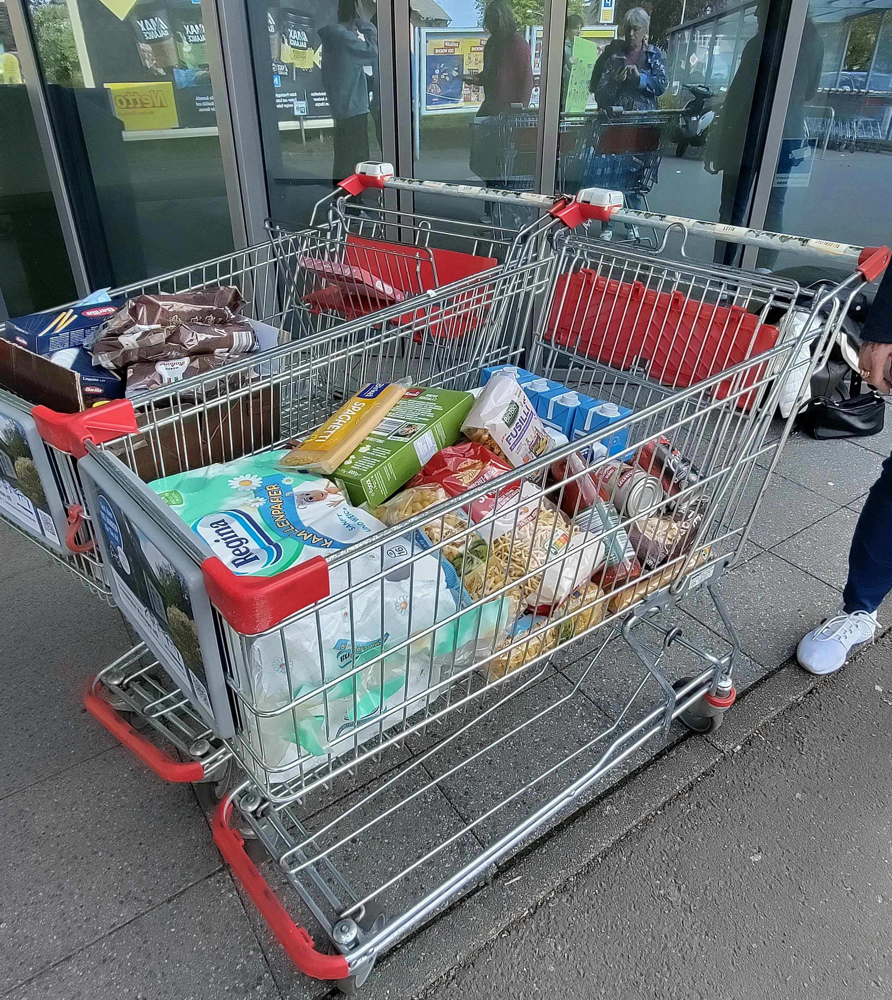 Ein voll beladener Einkaufswagen mit Lebensmitteln und Hygieneartikeln steht vor dem Eingang eines Supermarktes.