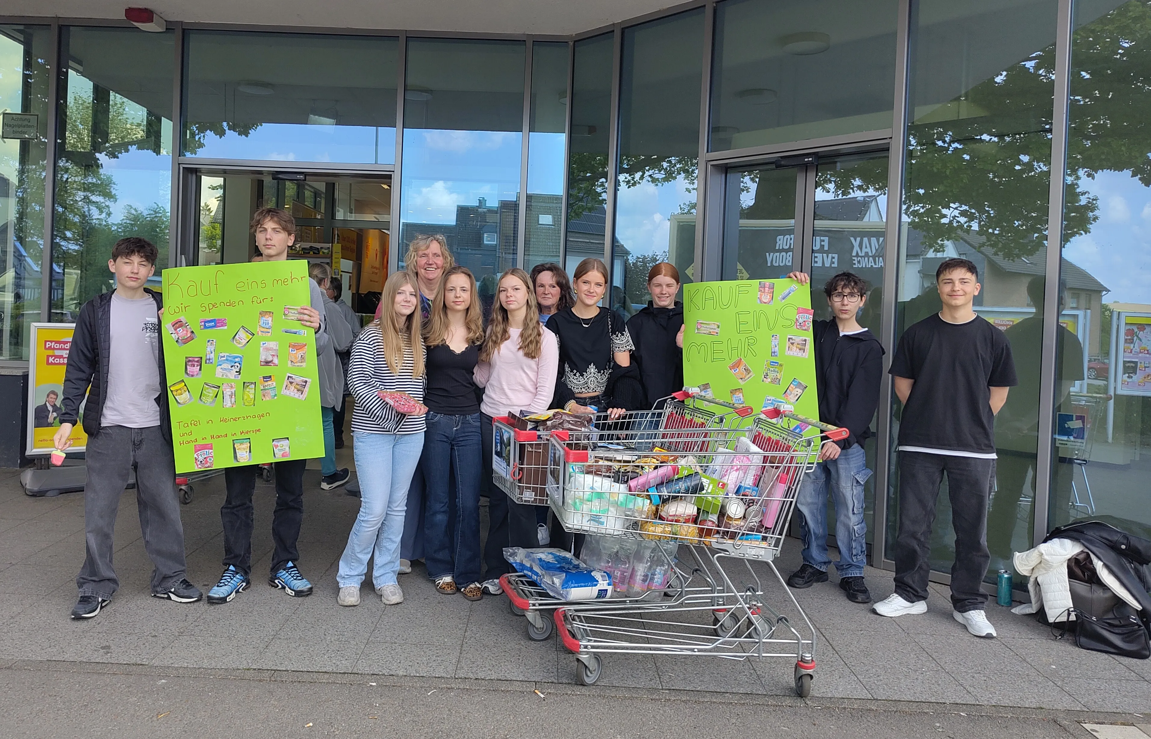 Gruppe Jugendlicher mit einem vollgepackten Einkaufswagen vor einem Supermarkt, sie halten Schilder mit der Aufschrift „Kauf eins mehr“.