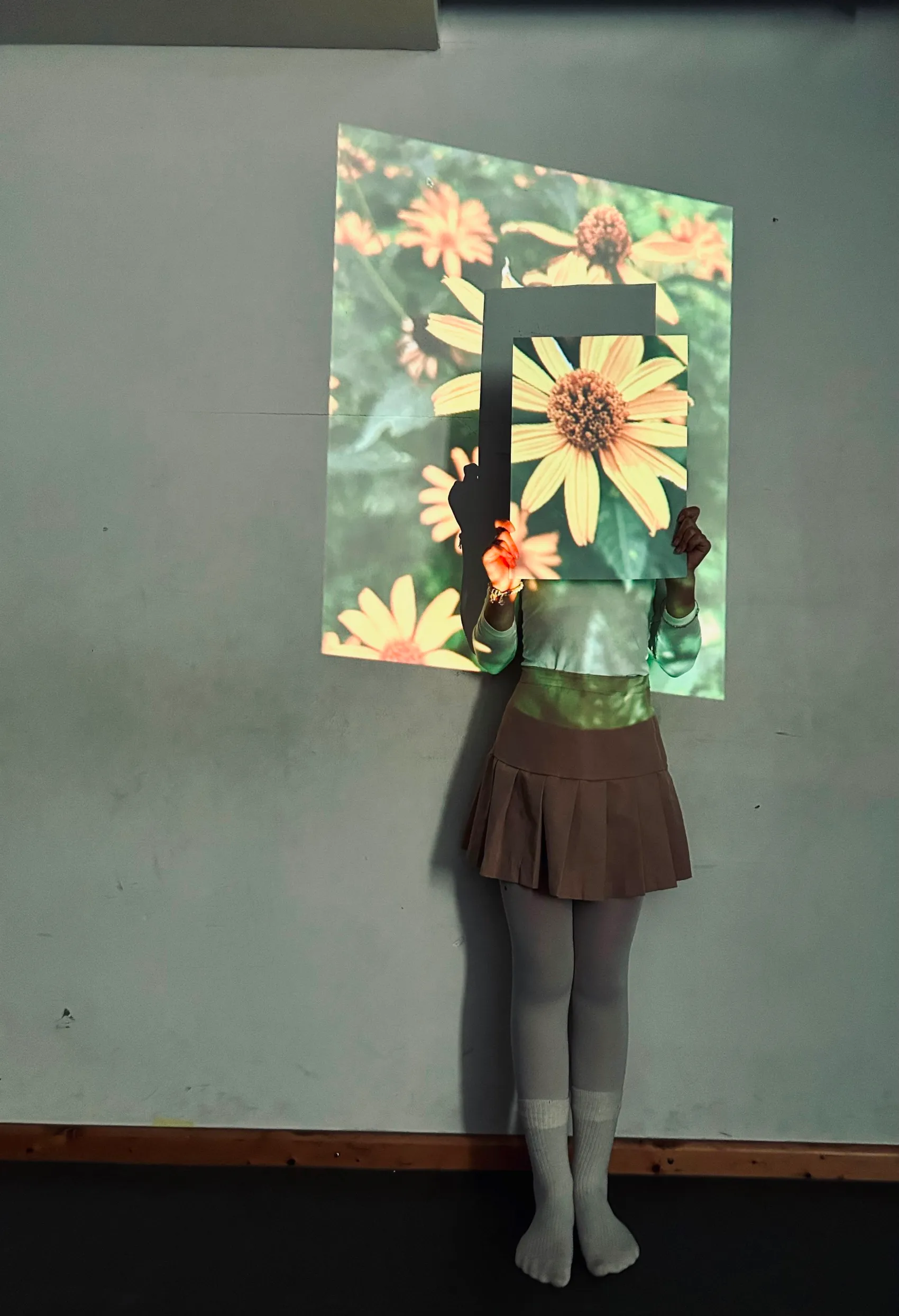 Ein Kind steht vor einer Projektion von Blumen und hält einen schwarzen Rahmen vor sein Gesicht, der die Form einer großen Blüte hat.