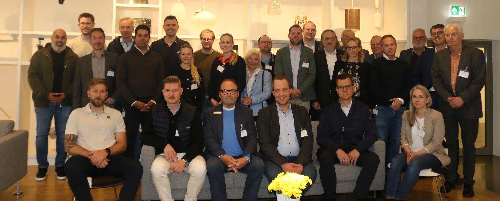 Gruppe von etwa dreißig Berufstätigen posiert für ein Gruppenfoto in einem hellen Innenraum. Im Vordergrund hält eine Person einen Strauß gelber Tulpen.