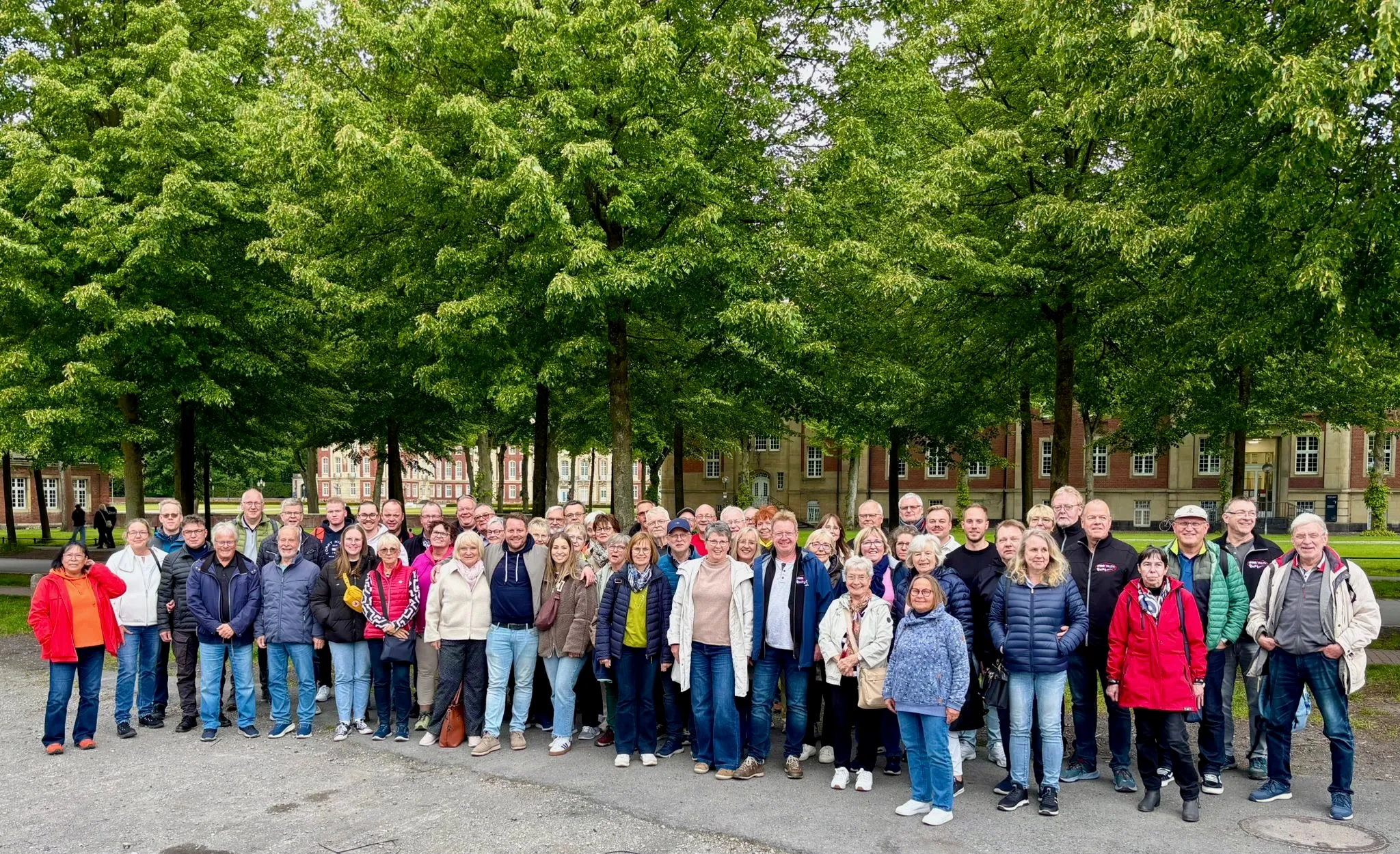 Eine große Gruppe von Menschen steht vor einem klassizistischen Schlossgebäude mit grünen Bäumen im Hintergrund für ein Gruppenfoto zusammen.
