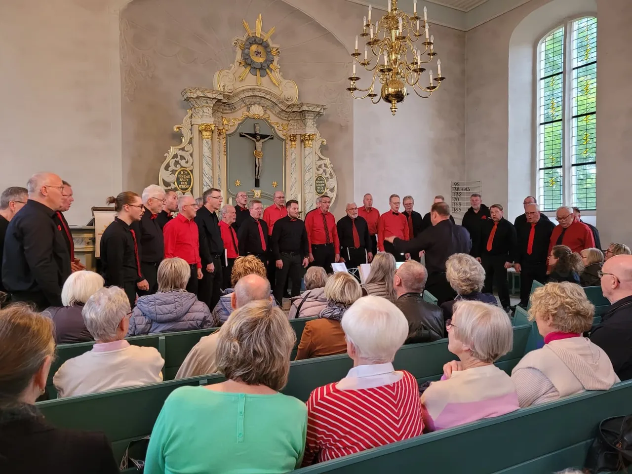 Chor Four Valleys singt in einer Kirche vor einem Publikum. Im Hintergrund ein Altar mit Kruzifix und eine Empore.