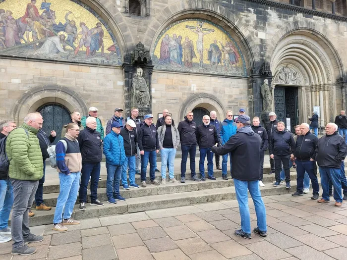 Eine Gruppe von Männern in Freizeitkleidung steht vor dem Bremer Dom und lauscht einem Mann, der gestikulierend etwas erklärt. Im Hintergrund ist die Fassade des Doms mit ihren kunstvollen Mosaiken zu sehen.