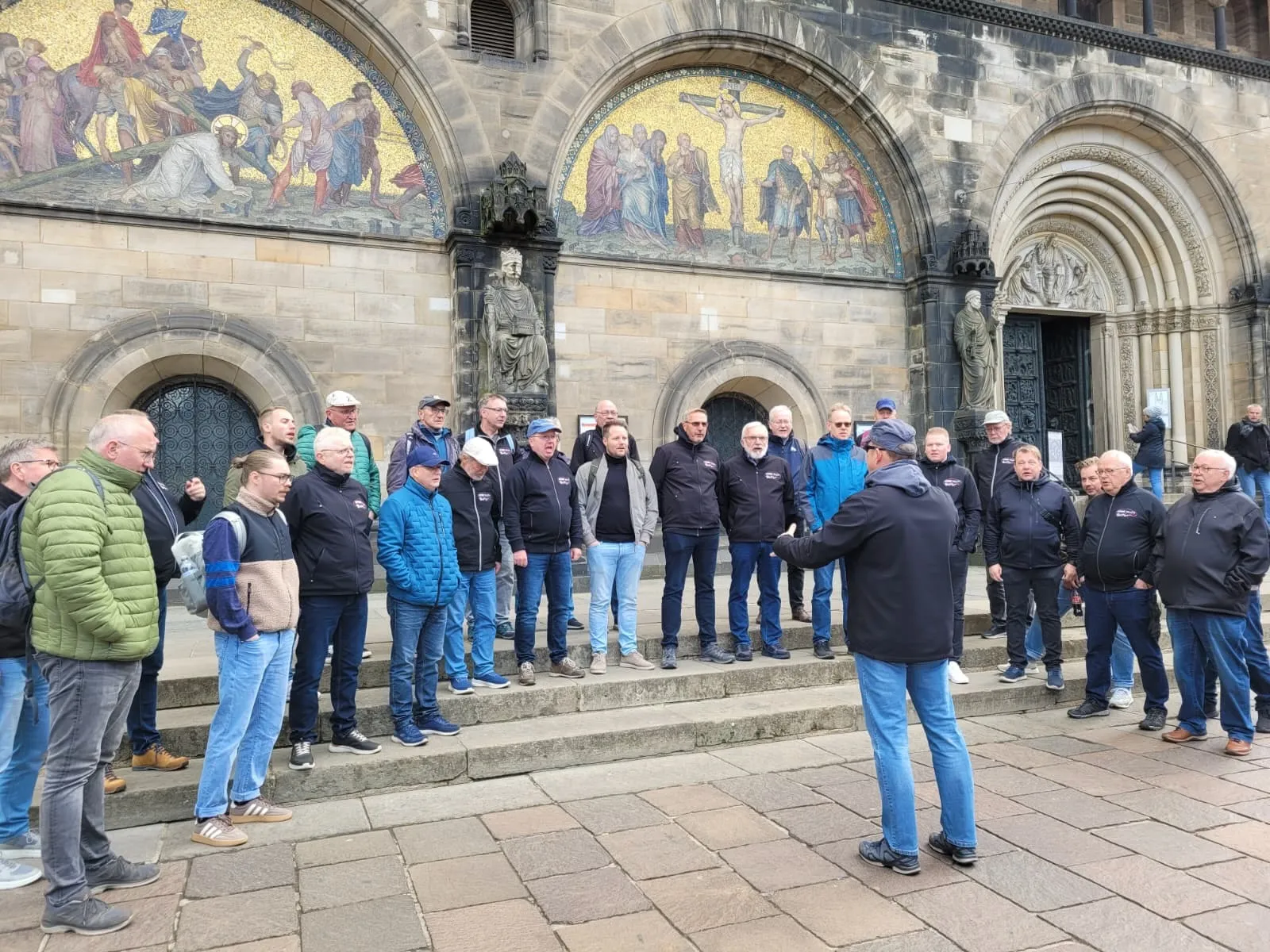 Eine Gruppe von Männern in Freizeitkleidung steht vor dem Bremer Dom und lauscht einem Mann, der gestikulierend etwas erklärt. Im Hintergrund ist die Fassade des Doms mit ihren kunstvollen Mosaiken zu sehen.