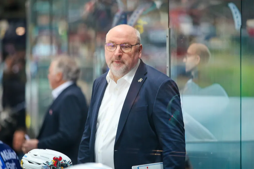 Frank Fischöder, ein Mann mit Brille und Bart, trägt Anzug und Hemd und blickt in die Ferne. Im Hintergrund ist eine Eishockeyarena zu sehen.
