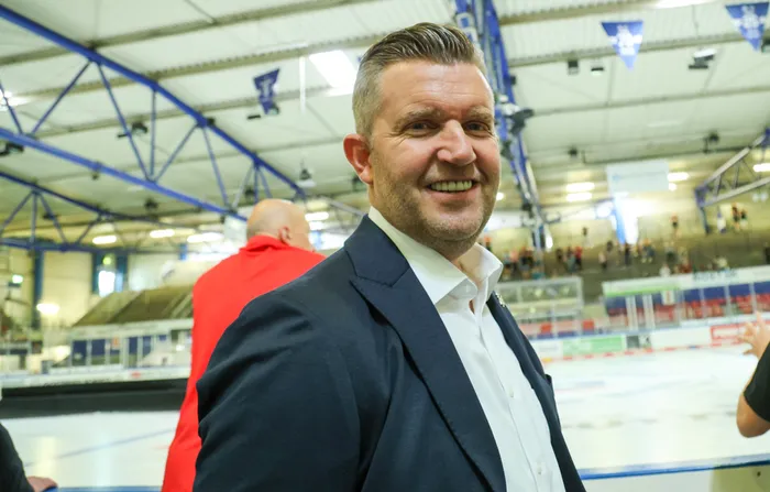 Sportdirektor Franz-David Fritzmeier lächelt in einer Eishalle vor einer Eisfläche.