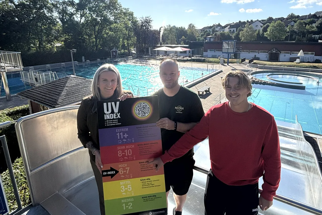 Drei Personen halten eine farbige UV-Index-Tafel vor einem Freibad.