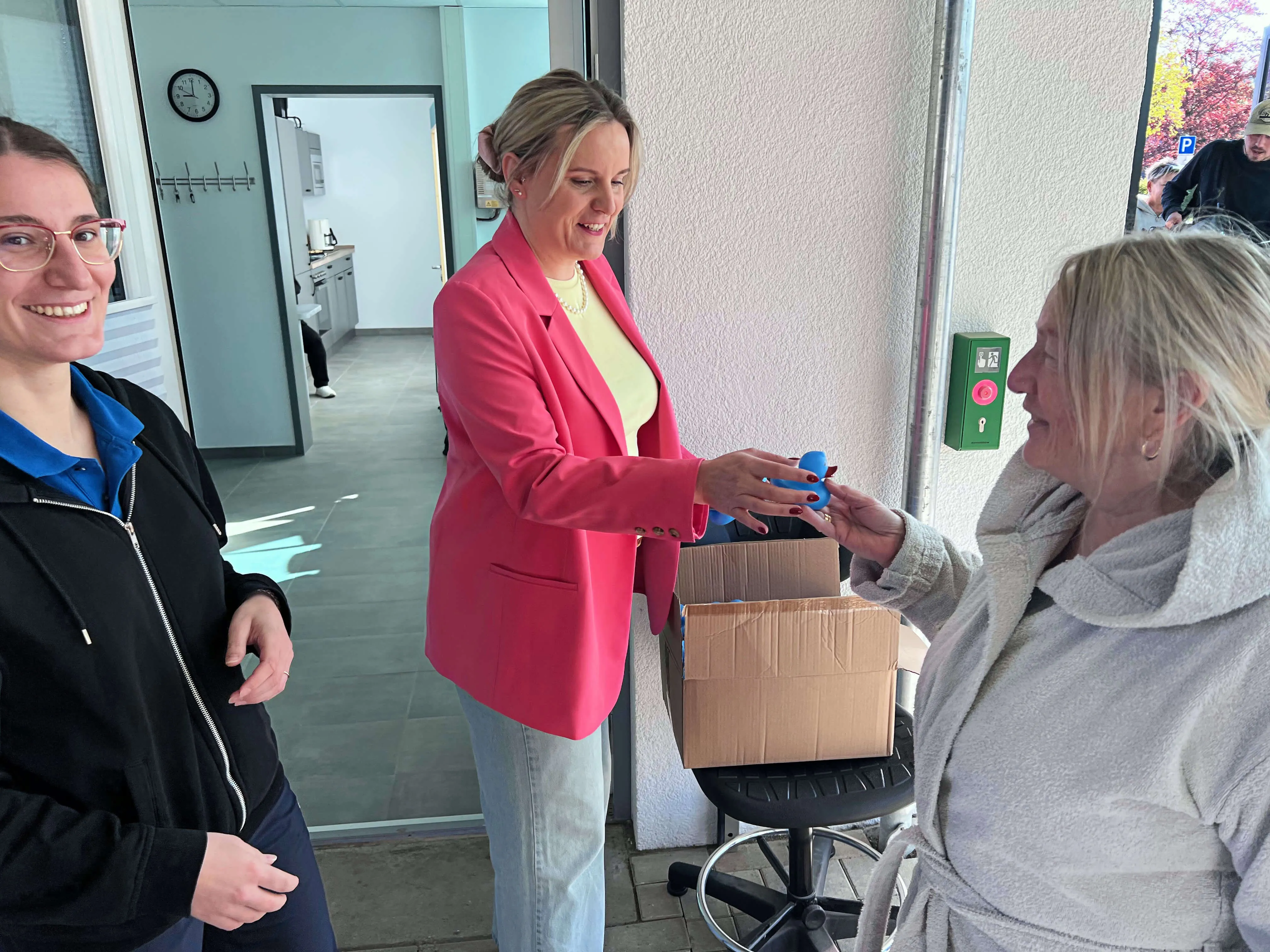 Frau in pinkem Blazer überreicht einer anderen Frau ein blaues Geschenk, während eine dritte Person lächelnd im Hintergrund steht.
