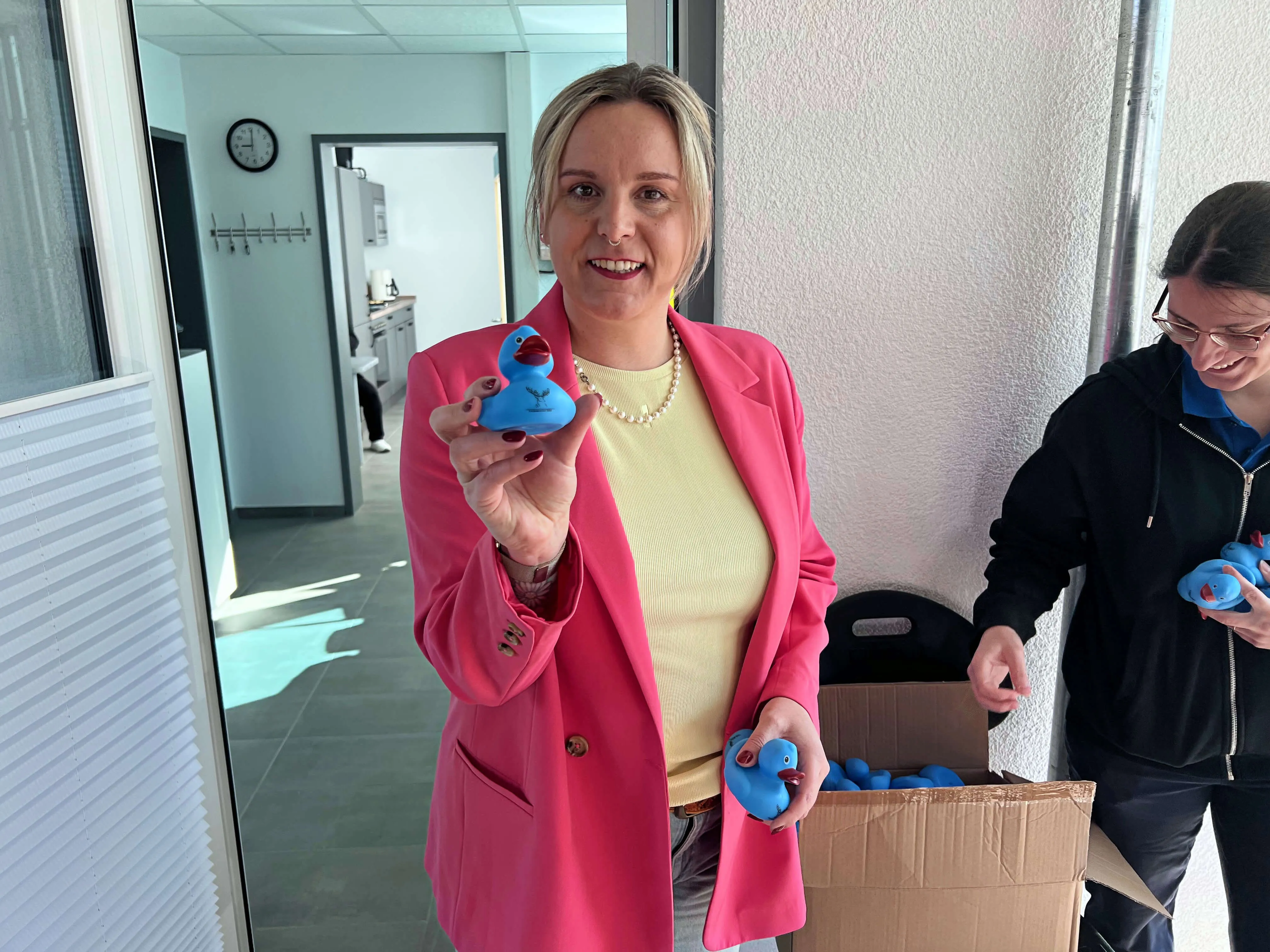 Frau in pinkem Blazer hält blaue Gummienten vor sich, während eine Kollegin im Hintergrund weitere Enten aus einem Karton nimmt. Im Hintergrund ist ein Büro zu sehen.