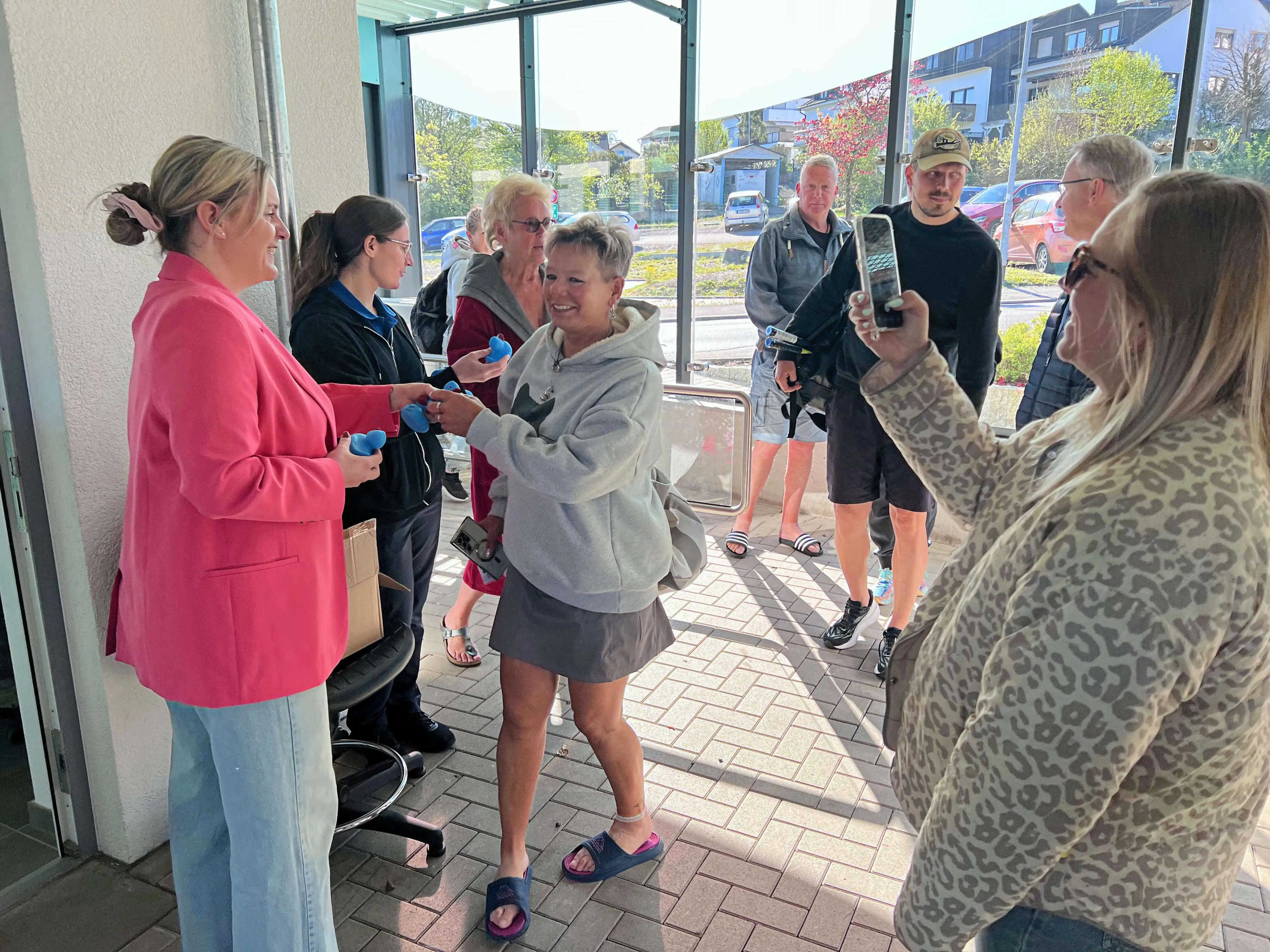 Frau in Jogginghose und Shorts hält zwei blaue Entenfiguren vor einer Gruppe von Menschen am Eingang des Freibads Herscheid.