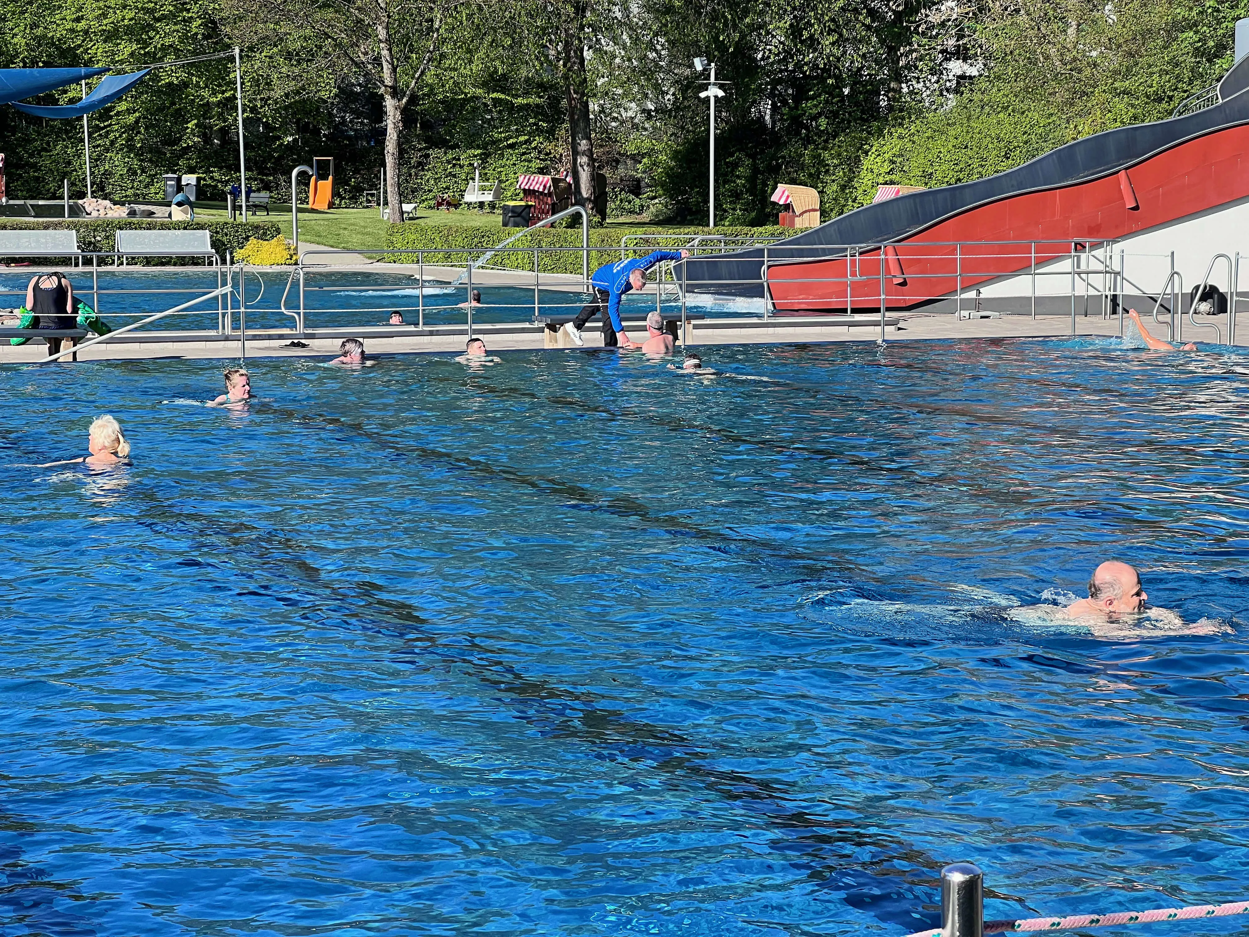 Menschen schwimmen und planschen im blauen Wasser des Freibads Herscheid, während andere am Beckenrand stehen oder sitzen. Im Hintergrund eine rote Wasserrutsche und Bäume.