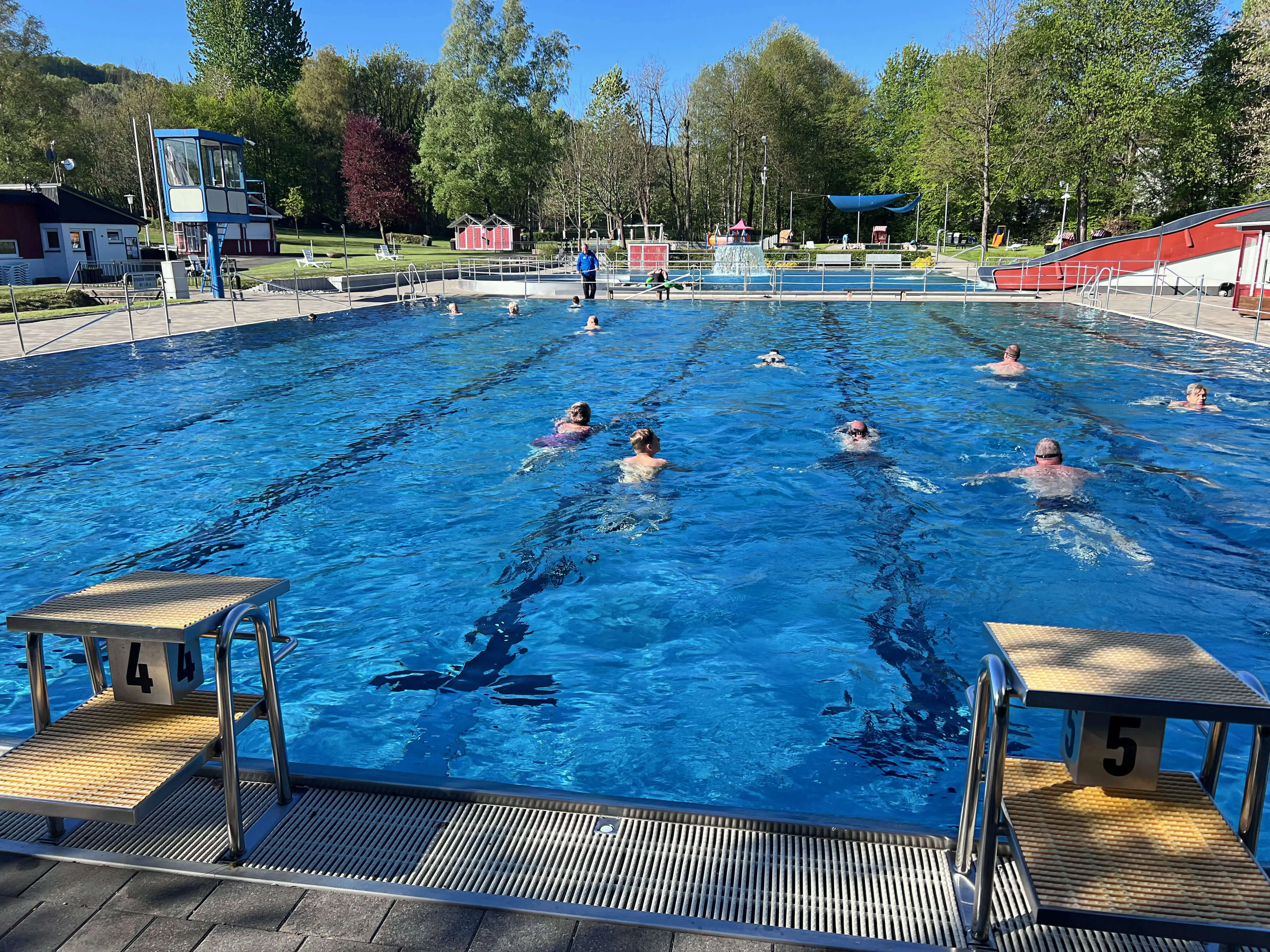 Mehrere Schwimmer in den Bahnen eines Freibads an einem sonnigen Tag. Im Vordergrund Startblöcke mit den Nummern 4 und 5.