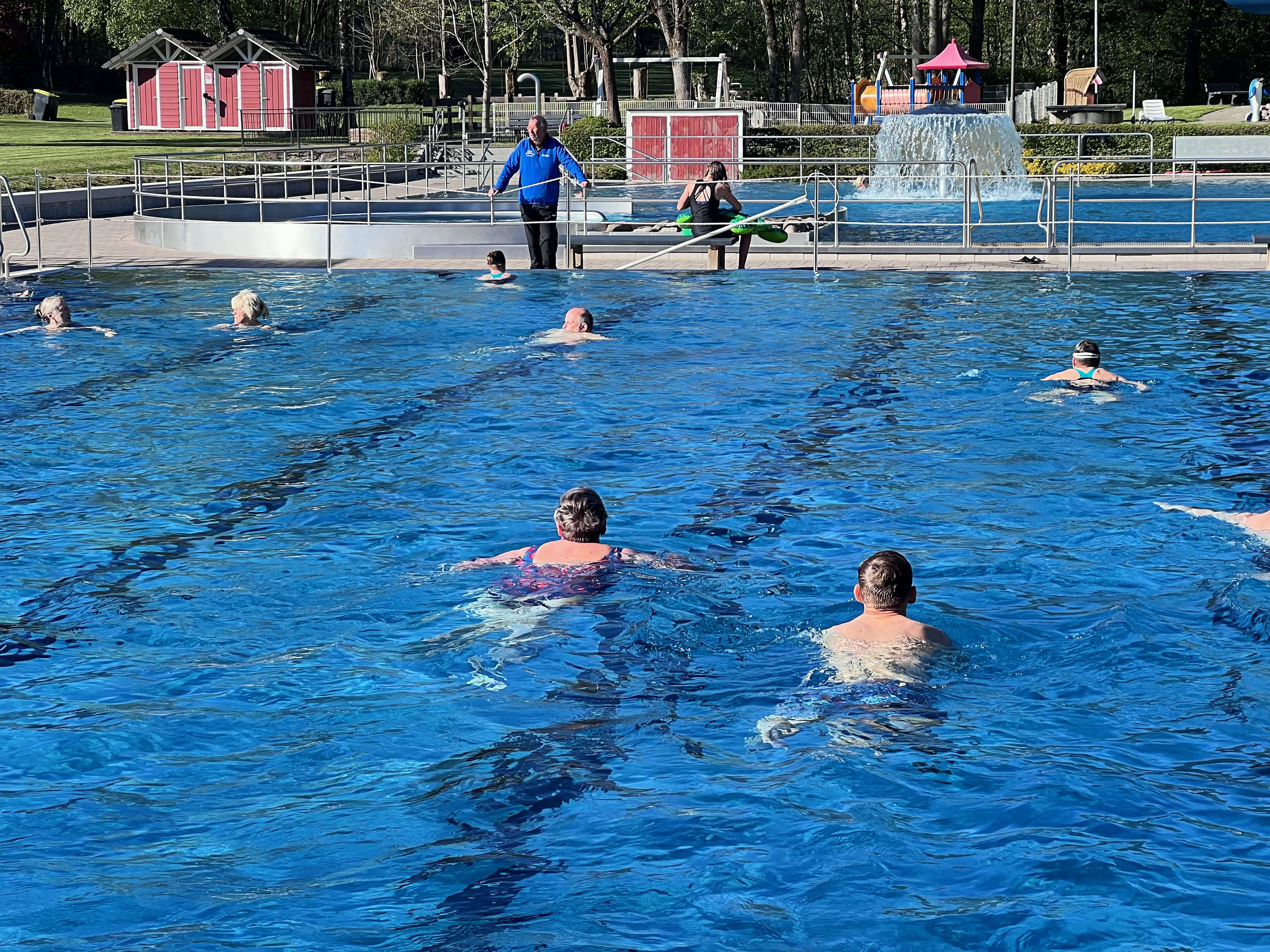 Menschen schwimmen im Freibad Herscheid, während ein Betreuer am Beckenrand steht und Anweisungen gibt.