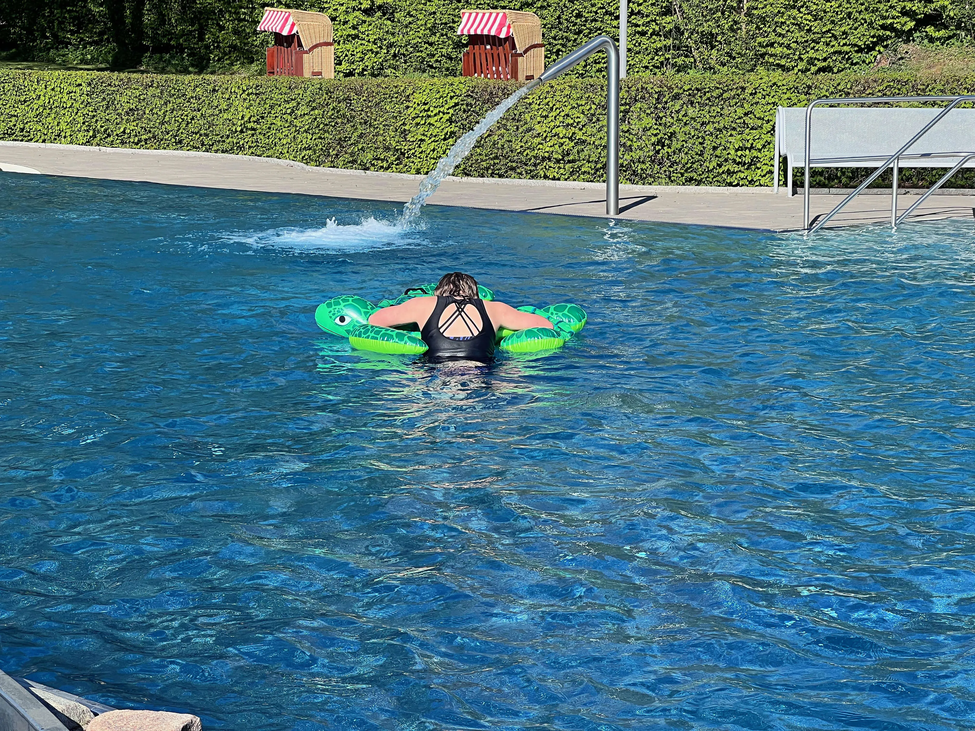 Eine Person schwimmt auf einem aufblasbaren grünen Frosch im blauen Wasser des Freibads Herscheid, während ein Wasserstrahl im Hintergrund sprüht.