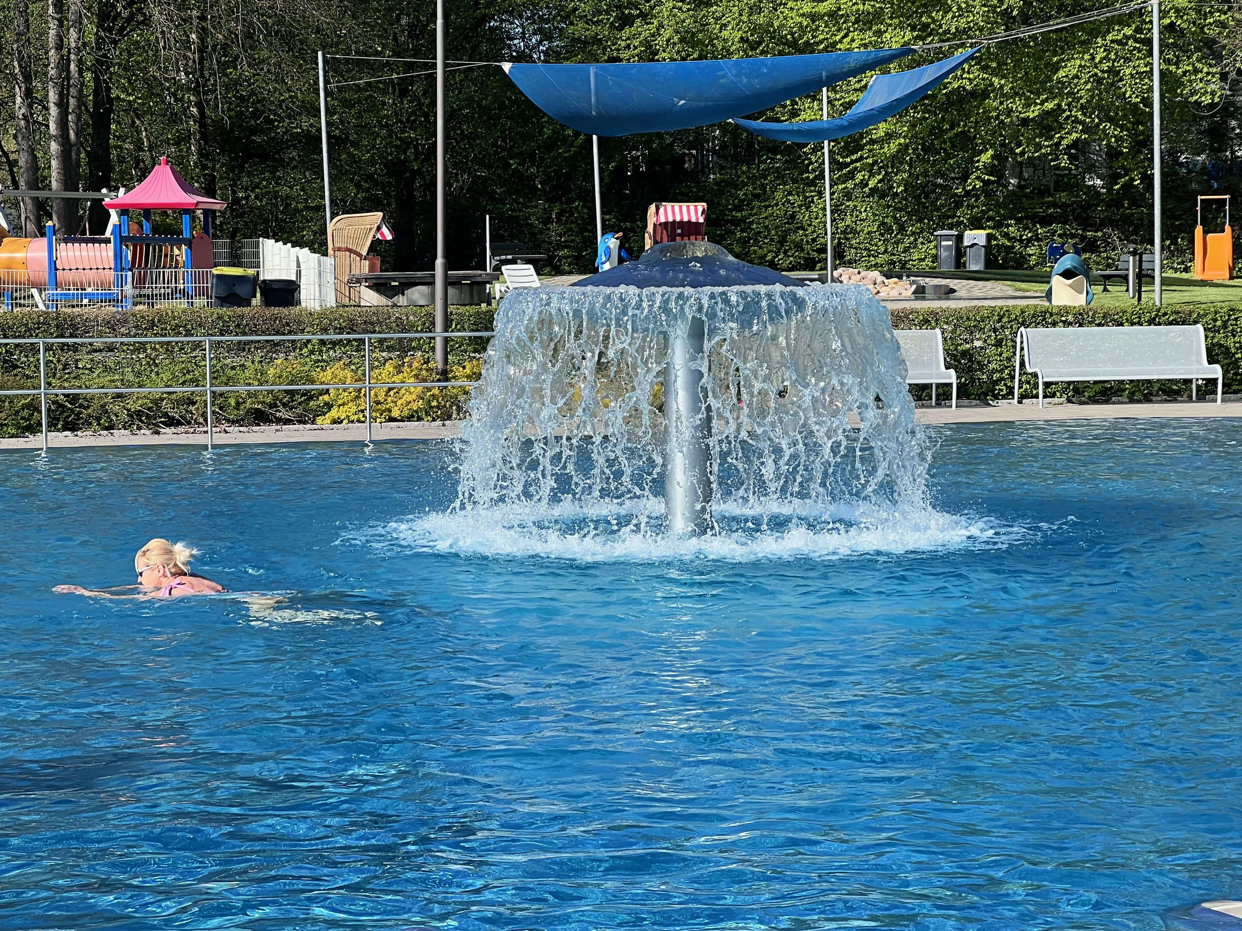 Ein Schwimmer im Freibad Herscheid unter einem Wasserfall, im Hintergrund ein Spielbereich mit Sonnensegel.