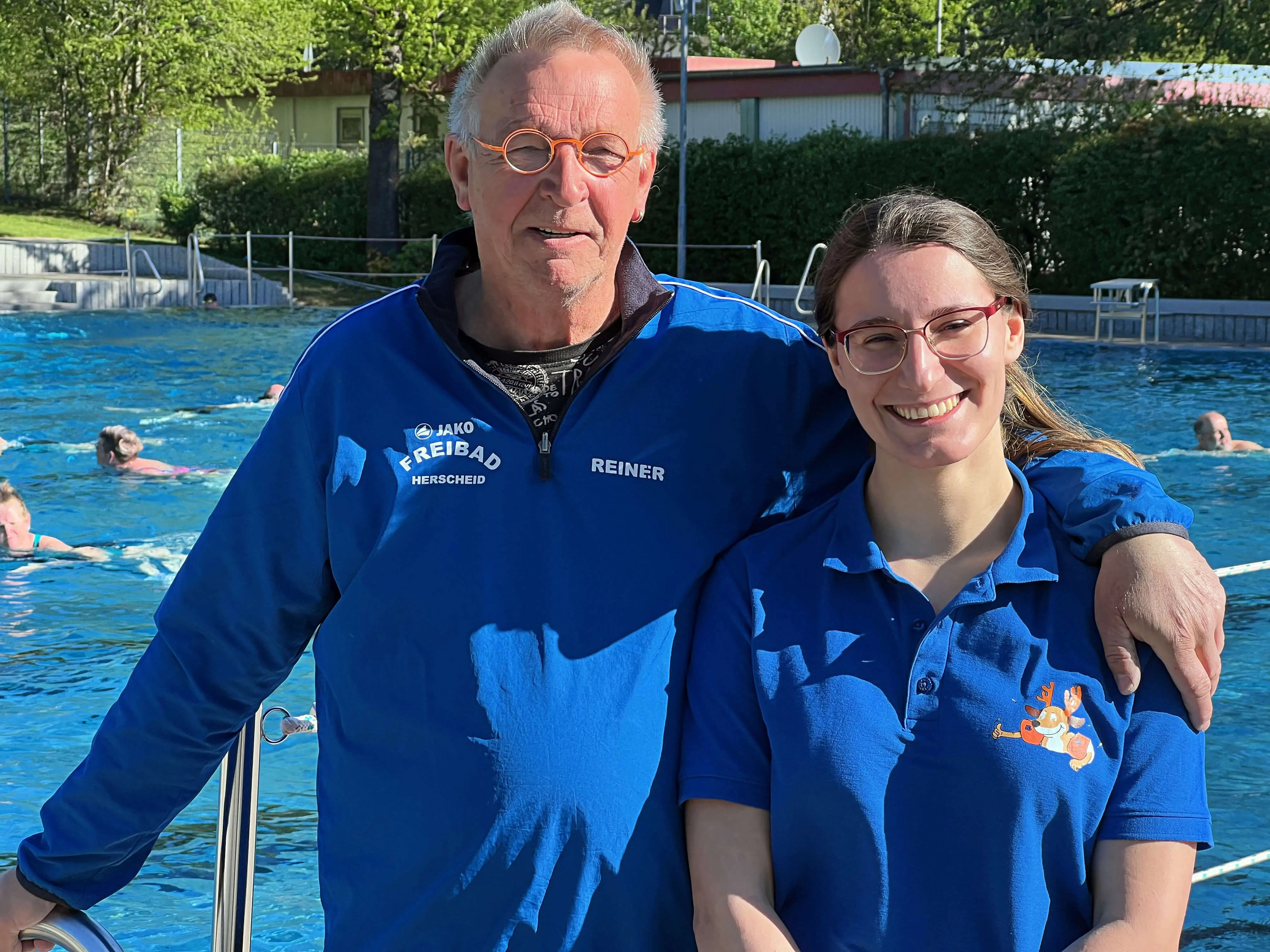 Ein Mann und eine Frau lächeln in die Kamera, im Hintergrund ein Schwimmbecken mit Badegästen. Der Mann trägt eine Jacke mit dem Aufdruck „JAKO FREIBAD HERSCHEID“.