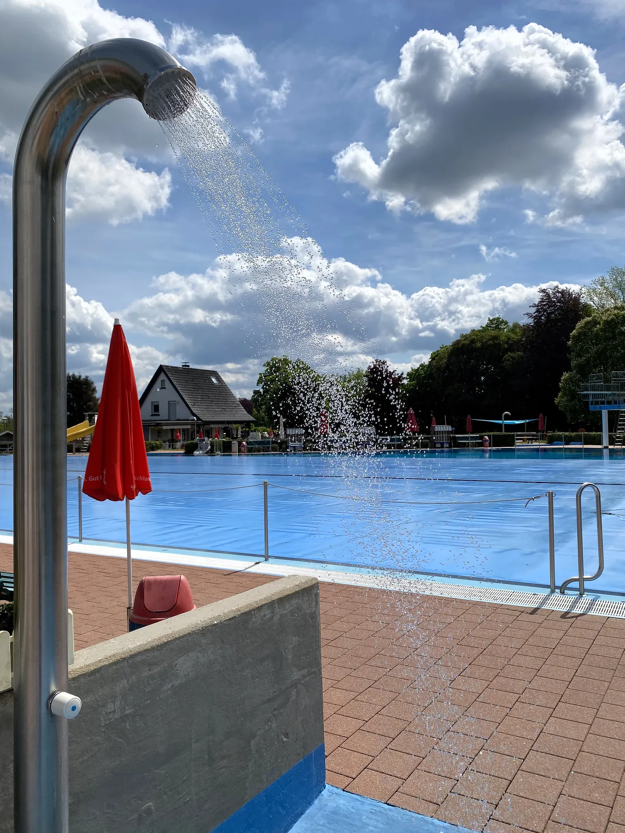 Eine Außendusche sprüht Wasser über das Becken eines Freibads an einem sonnigen Tag. Im Hintergrund sind Bäume und Gebäude zu sehen.