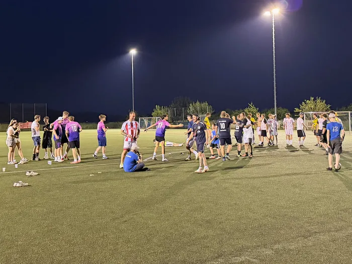 Eine Gruppe von Fußballspielern steht auf einem Kunstrasenplatz im Dunkeln zusammen, möglicherweise nach einem Spiel. Im Hintergrund sind Flutlichtmasten und Zuschauer zu sehen.