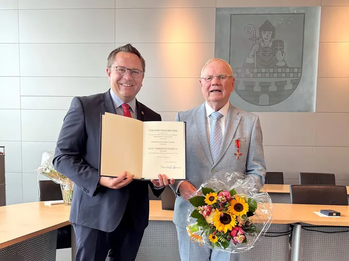 Marco Voge überreicht Friedrich-Karl Schmidt eine Urkunde. Schmidt hält einen Blumenstrauß und trägt das Bundesverdienstkreuz am Revers.