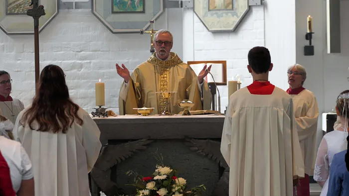 Ein Priester mit Brille und goldenem Gewand zelebriert einen Gottesdienst vor einem Altar mit Kerzen und sakralen Gegenständen. Mehrere Personen in weißen Gewändern stehen dem Priester zugewandt.