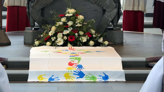 Ein weißer Tisch mit bunten Handabdrücken steht vor einem Blumenarrangement und einer steinernen Figur in einer Kirche.
