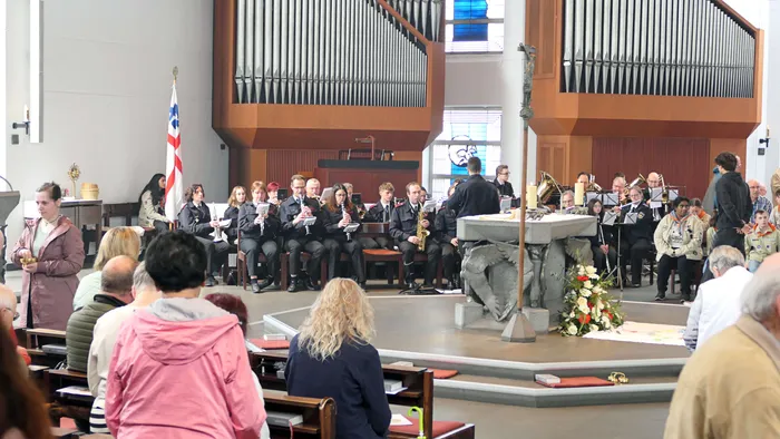 Blasmusik spielt während eines Gottesdienstes in der Kirche St. Laurentius. Im Vordergrund sitzen und stehen Besucher, während der Altar mit Blumen geschmückt ist.