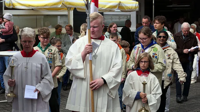 Eine Prozession mit Geistlichen und Jugendlichen zieht vor Zuschauern durch eine Stadt. Vorne trägt ein Mann in priesterlicher Kleidung ein Banner, während andere Teilnehmer Kerzen und Kreuze tragen.