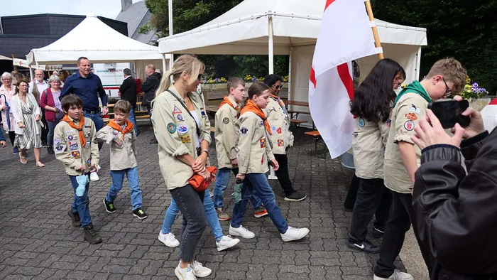 Eine Gruppe von Pfadfindern und Erwachsenen schreitet in Uniformen während einer Prozession vor einer Menschenmenge und weißen Zelten. Eine Person filmt die Prozession mit einer Kamera.