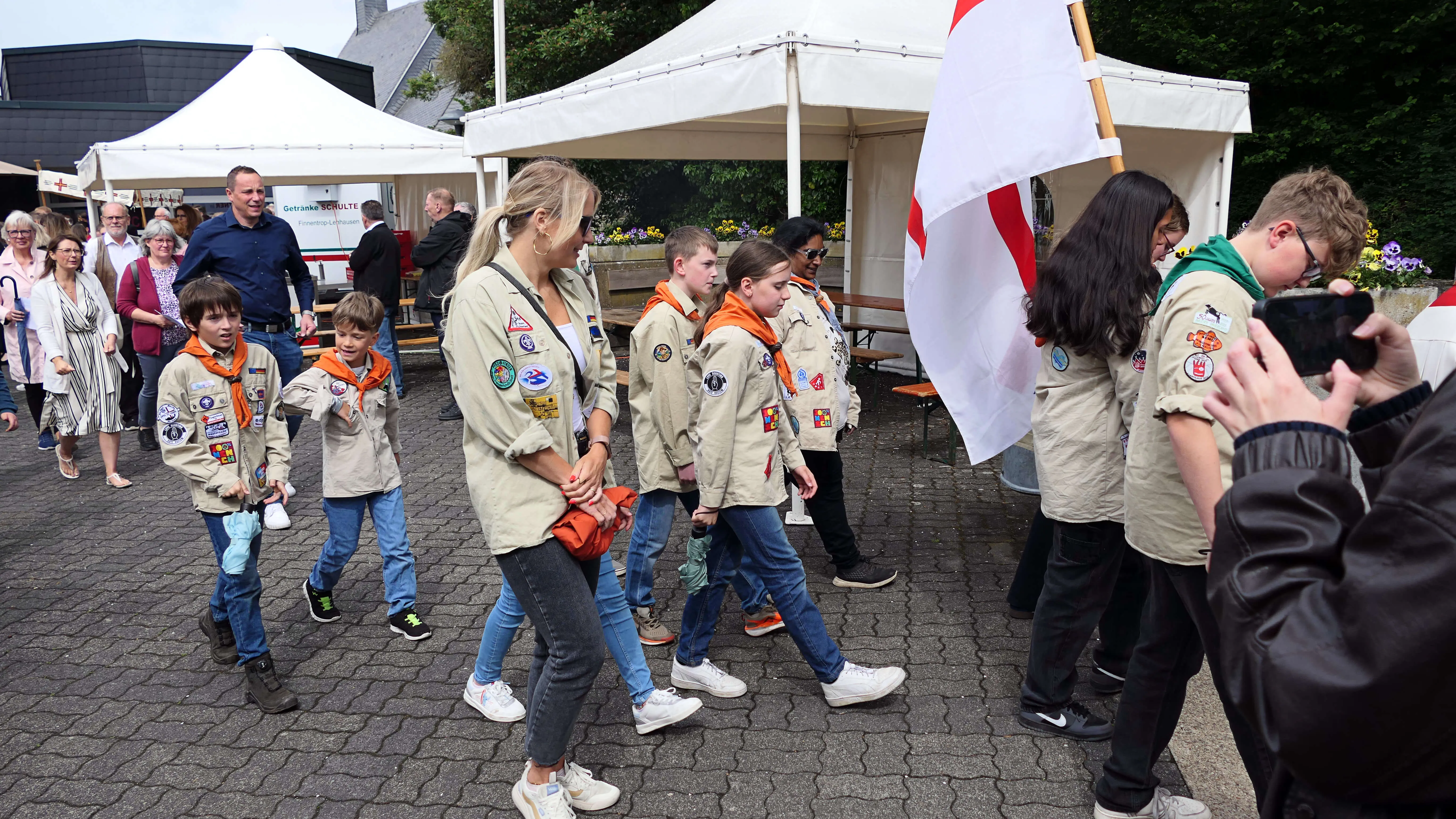 Eine Gruppe von Pfadfindern und Erwachsenen schreitet in Uniformen während einer Prozession vor einer Menschenmenge und weißen Zelten. Eine Person filmt die Prozession mit einer Kamera.
