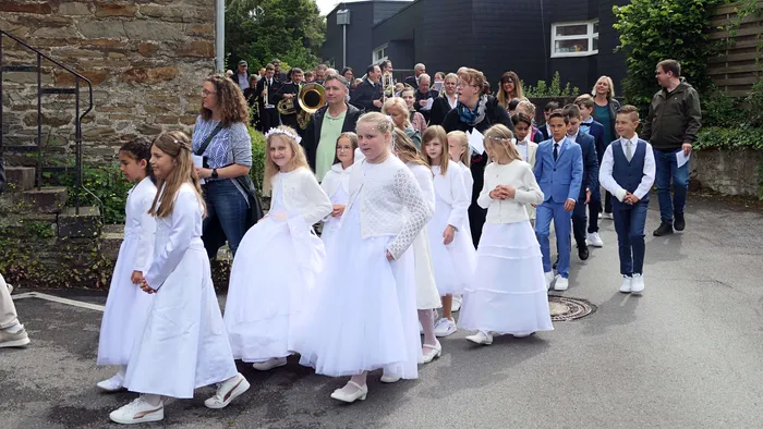 Kinder in weißen Gewändern ziehen in einer Prozession auf einer Straße, begleitet von einer Blaskapelle und zahlreichen Zuschauern.